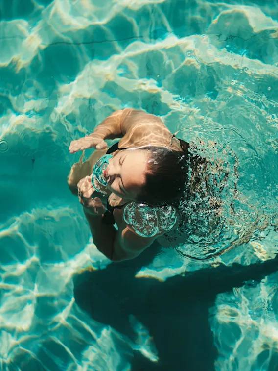 En person svømmer gledelig i et klart blått basseng, med vannsprut og bobler rundt ansiktet, og nyter et forfriskende øyeblikk.