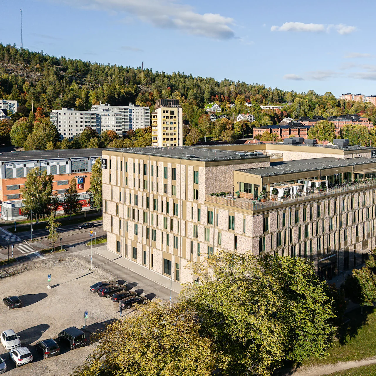 En fleretagers bygning med en tagterrasse, beliggende i et boligområde omgivet af grønne omgivelser og boliger. Parkerede biler og veje ses tæt ved under en klar himmel.