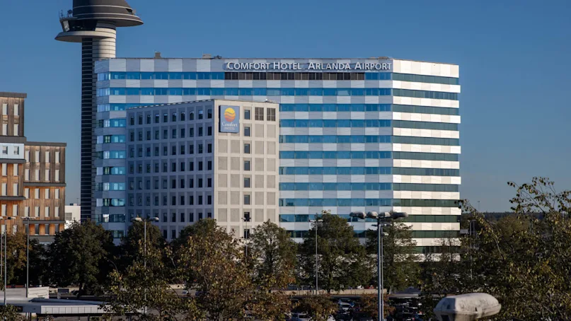 A large, modern hotel building labeled "Comfort Hotel Arlanda Airport" with reflective windows stands beside an airport control tower under a clear blue sky. Trees and other buildings surround the foreground.