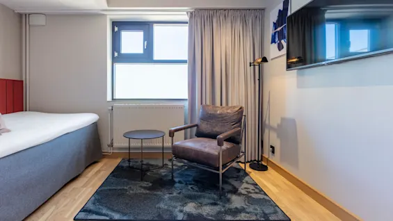 A modern hotel room with a bed, armchair, small table, and flat-screen TV on the wall.