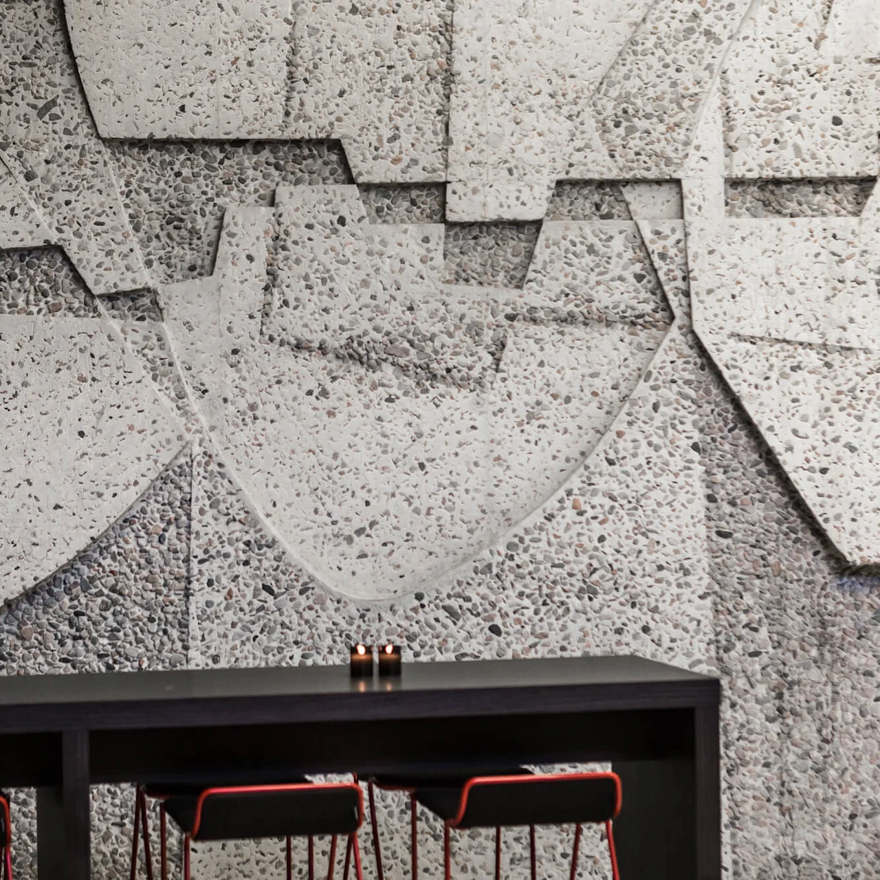 A black table with candles sits in front of a textured, geometric stone wall. Two chairs with red accents are tucked under the table.