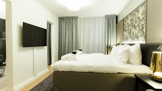 A neatly made bed with white linens rests in a modern hotel room. A flat-screen TV is mounted on the wall to the left. Curtains cover a large window.