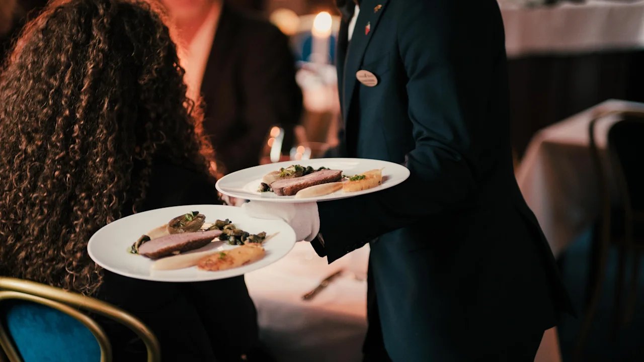 En servitör i mörk kostym serverar eleganta middagstallrikar till gäster vid ett restaurangbord.