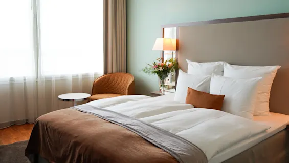 Bed, neatly made with brown and white linens, sits beside a bedside table with a lamp and vase of flowers, next to a window with sheer curtains and an armchair.