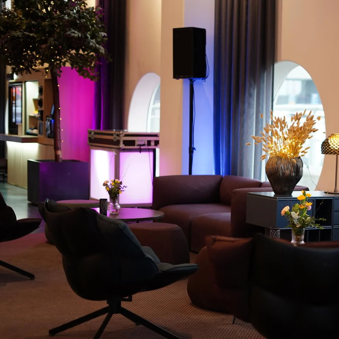 En siddeområde har moderne, mørke møbler med runde borde med blomsterarrangementer. Placeret nær store cirkulære vinduer skaber stemningsbelysning en farverig, indbydende atmosfære.