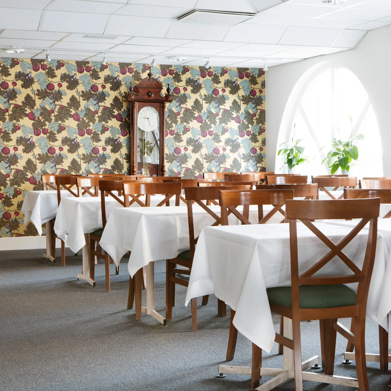 Wooden tables with white tablecloths in a dining room, sunlight entering through arched windows, a patterned wallpaper and a grandfather clock.