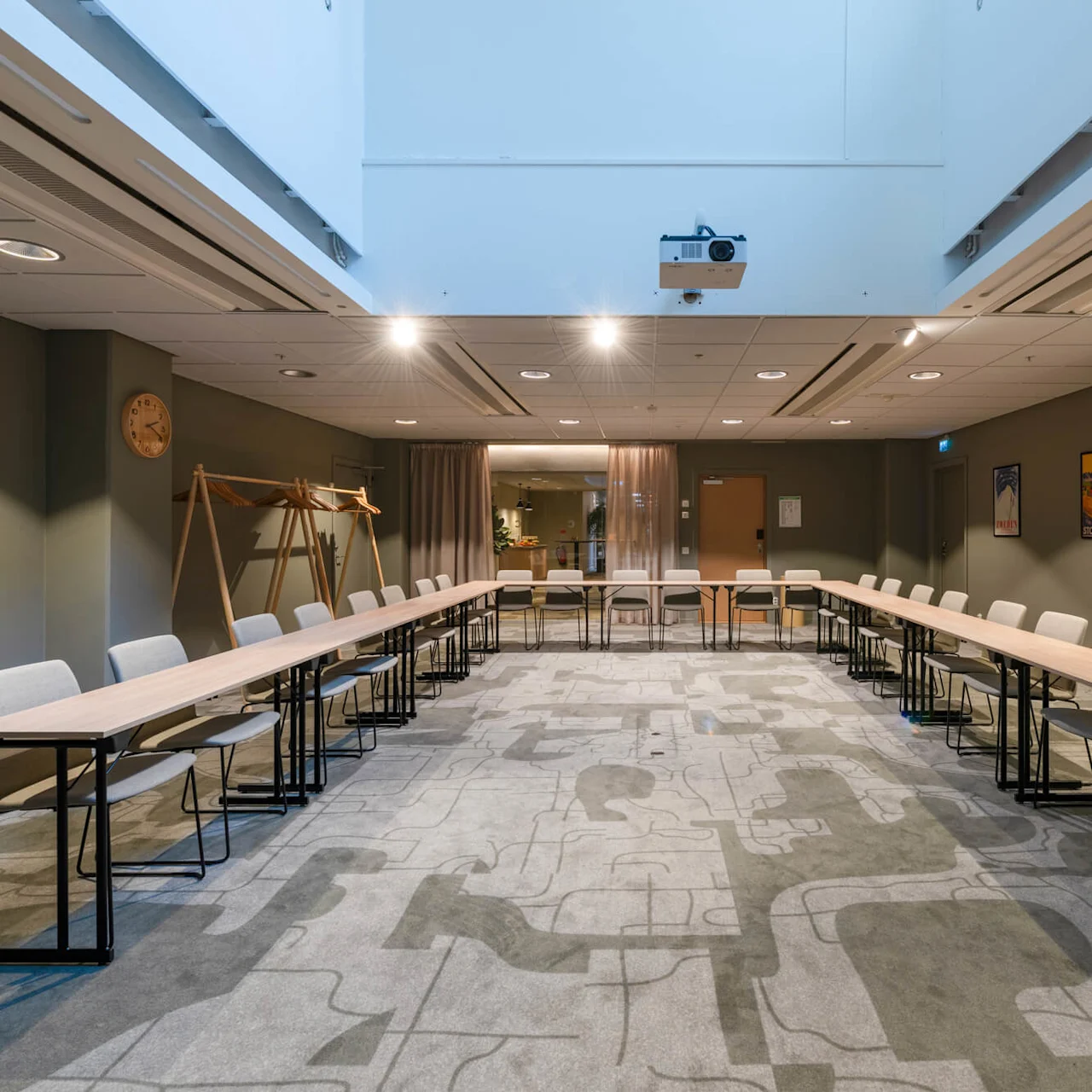 Meeting room with U-shaped table arrangement, surrounded by chairs. A projector is mounted on the ceiling. Walls have posters, a clock, and a whiteboard, and light beige carpet covers the floor.