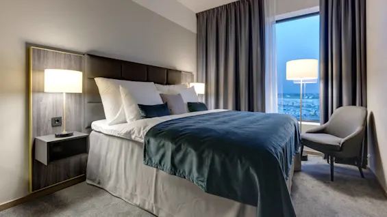 Seng med turkis sengetøy og hvite puter står mellom to tente lamper i et moderne hotellrom, ved siden av et stort vindu med lukkede gardiner og en lenestol.