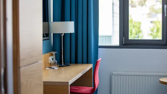 Et skrivebord av tre med en lampe og et glass står ved et vindu, omgitt av blå gardiner og en rød stol, i et lyst rom.