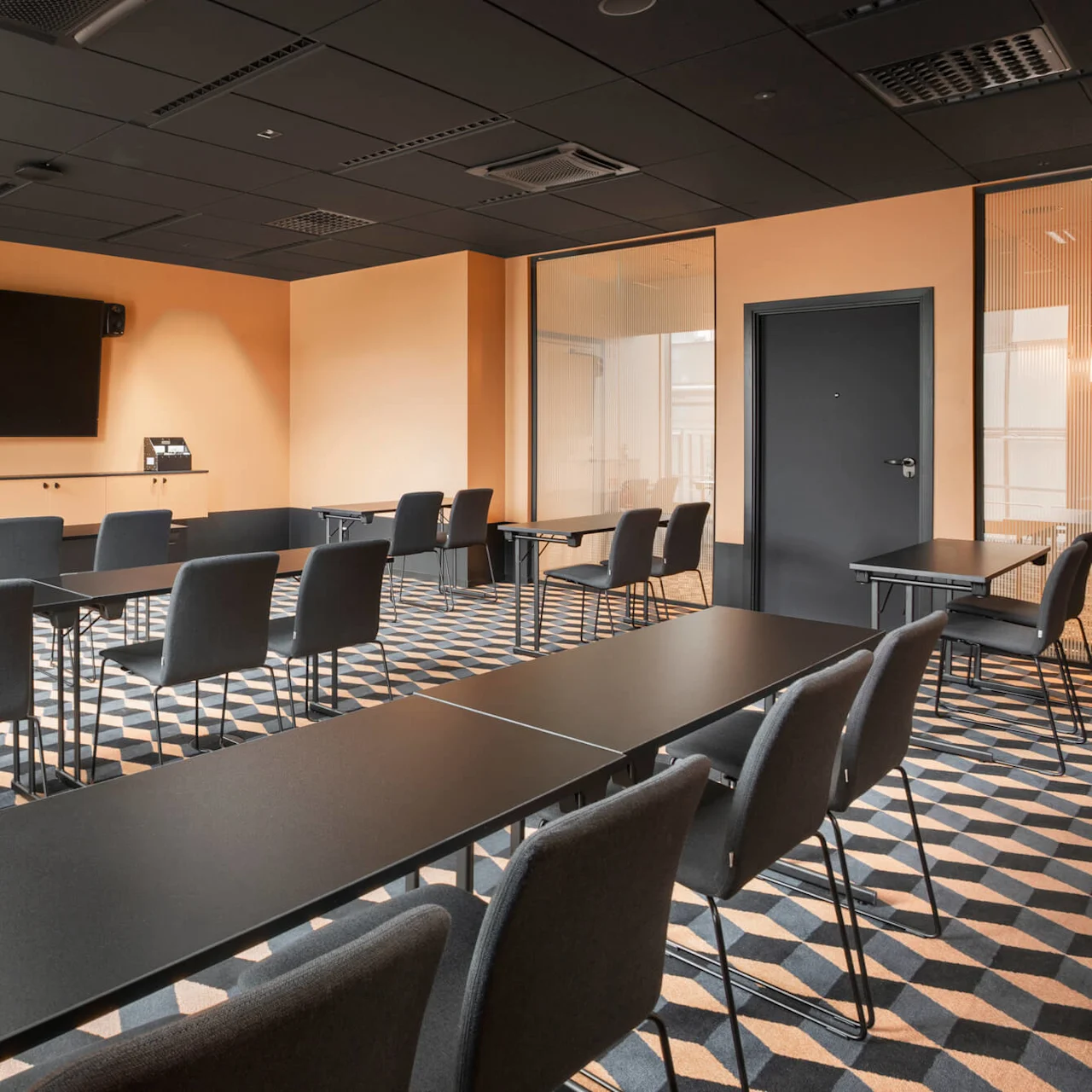 Ett konferensrum har rader av svarta stolar som vetter mot en stor väggmonterad skärm, mot orange väggar och ett mönstrat golv. Bord är arrangerade för en presentation.