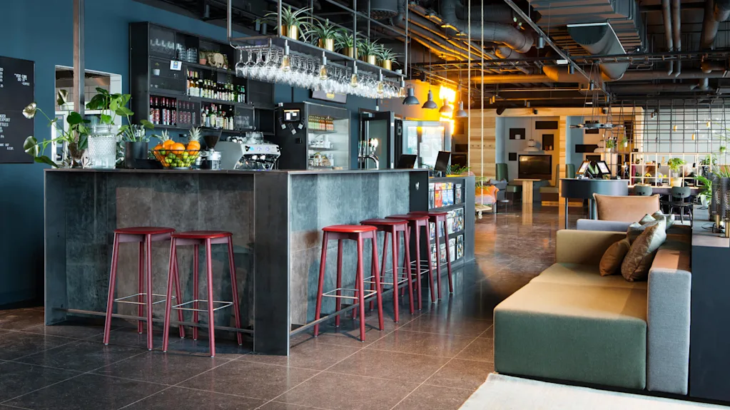 A modern bar area with stools and various seating arrangements in a spacious industrial-style lounge with exposed ductwork and warm lighting.