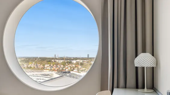Et rundt vindue tilbyder en panoramaudsigt over en bysilhuet under en klarblå himmel, med et skrivebord og en lampe i nærheden.