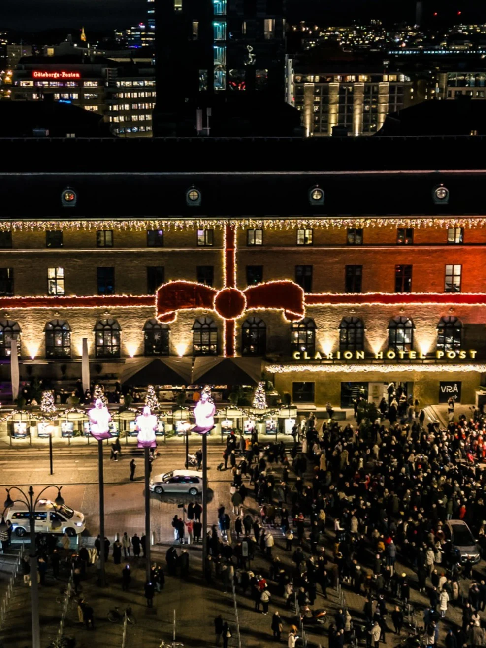 A large, historic building with bright decorative lights hosts a gathering of people at night, surrounded by city lights and nearby roads.