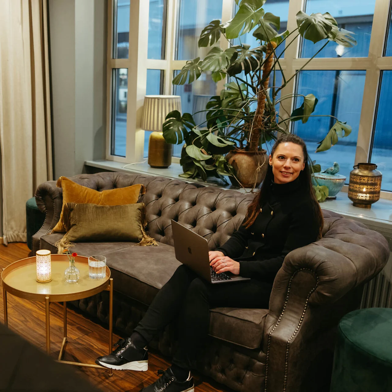 Nainen sohvalla käyttää kannettavaa tietokonetta viihtyisässä olohuoneessa, jossa on kasveja ja lämmintä valaistusta.