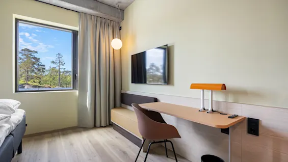A modern hotel room with a desk, TV, and a large window offering a serene view of lush trees and blue sky.