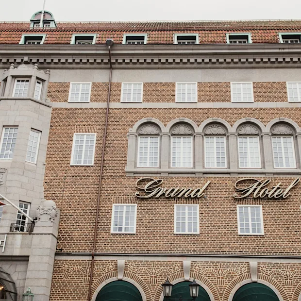 Den ståtliga tegelfasaden på ett hotell, med många fönster och en framträdande skylt där det står 'Grand Hotel'. Entrén säger 'GRAND'.