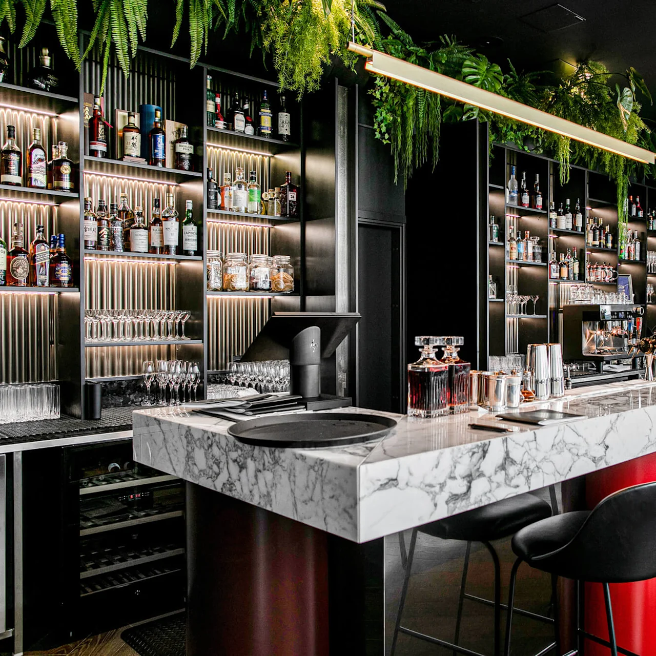 A marble bar counter with black chairs displays cocktail-making tools and a flower arrangement. Shelves behind hold various liquor bottles amid ambient lighting and lush hanging plants.