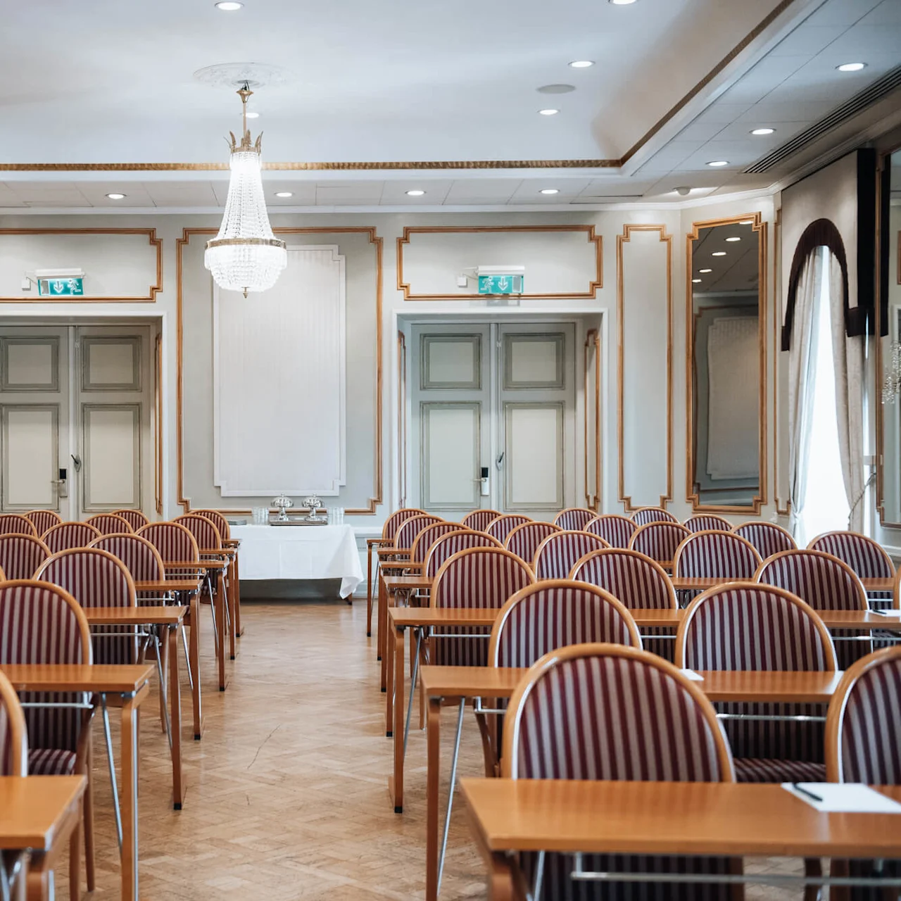 Konferencelokale med rækker af rødstribede stole og træborde, vendt mod en væg med dobbeltdøre. En lysekrone hænger fra loftet. Papirer ligger på bordene.