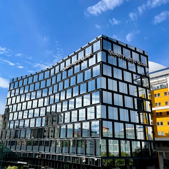 En modern glasbyggnad visar "QUALITY HOTEL GLOBE" på fasaden och speglar en solig blå himmel. Den står mot en stadsmiljö med närliggande gula och vita byggnader.