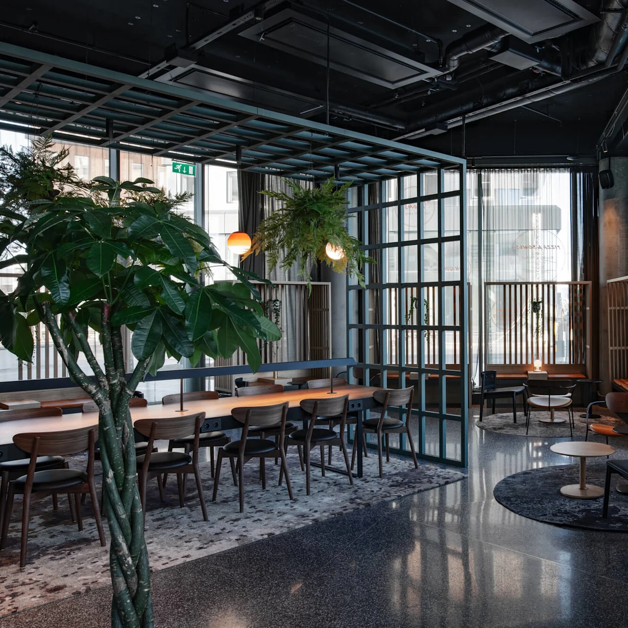 A spacious, modern lounge area features a large wooden table with chairs, surrounded by greenery and ambient hanging lights, creating a cozy, inviting atmosphere.