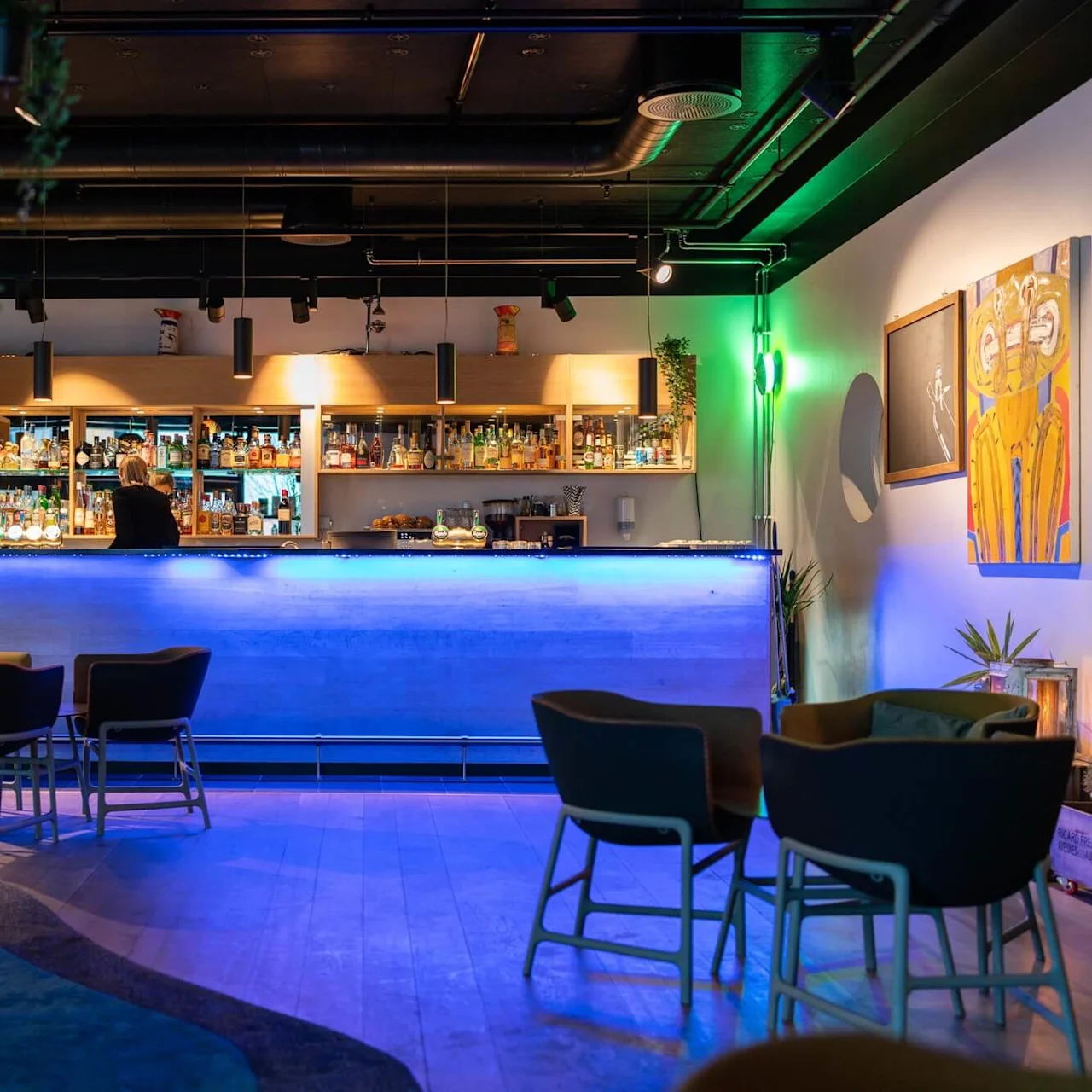 En modern bar med en blålysande disk och olika flaskor på hyllor. En person står bakom baren. Omkring står bord och stolar som skapar en mysig atmosfär.