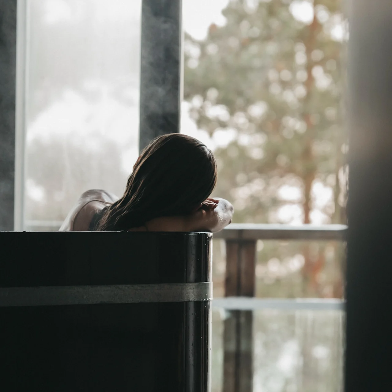 A person relaxing in a steaming hot tub, looking out a large window at trees.
