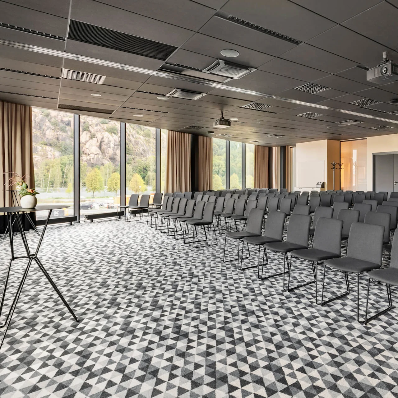 Rader av grå stolar arrangerade i ett konferensrum med fönster. Stora fönster visar en naturskön utsikt utomhus med träd och klippor, och geometriskt mönstrad matta täcker golvet.