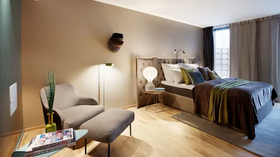 A modern bedroom features a bed with decorative pillows beside a round side table and lamp. A cushioned chair and footrest face a small table with books. Wooden floors create warmth.