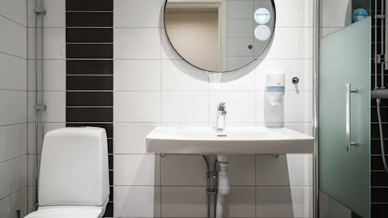 En vit badrumshandfat med kromkran monterad på en kaklad vägg, placerad under en rund spegel. Text på spegel: ”DRINK MORE WATER”. Närliggande toalett och dusch med frostad glasdörr.