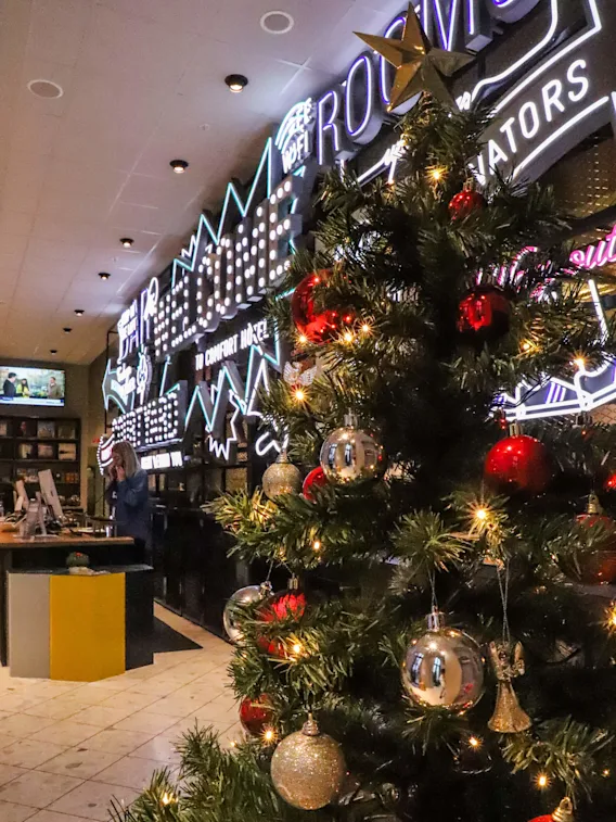 En dekorerad julgran med röda och silverfärgade prydnader står i en ljus lounge. Bakom finns en neonskylt med texten "ROOMS". Människor är vid receptionsdisken.