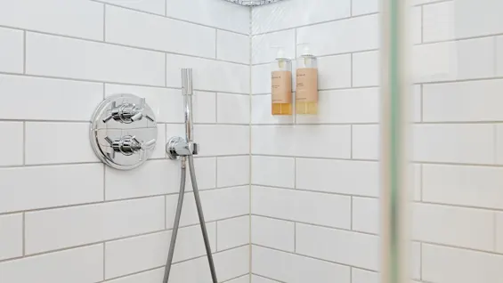 Et rent, moderne brusebad med hvide fliser, håndbruser og toiletartikler på en hjørnehylde.