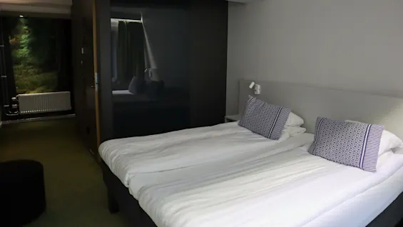 To senger med hvitt sengetøy står i et moderne hotellrom, med mønstrede puter og en svart skyvedør, ved siden av en gang med skogstapet.