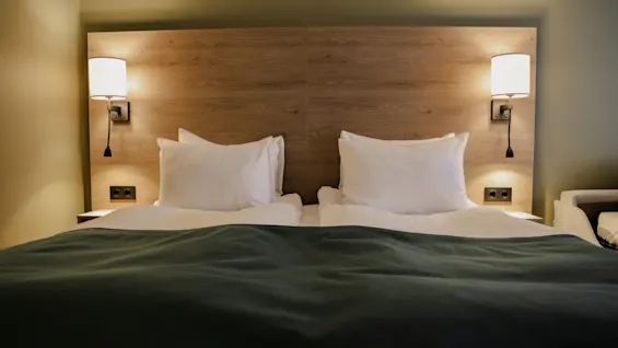 A bed with green blanket and white pillows rests under a wooden headboard, flanked by two wall-mounted lamps. Soft lighting highlights the neutral-colored room.