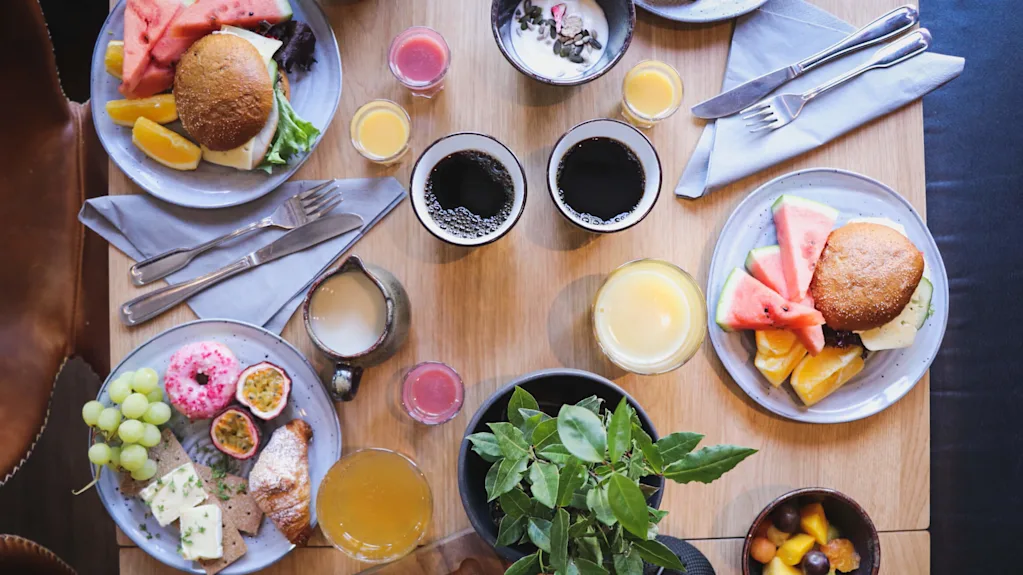 An overhead shot of a brunch spread with sandwiches, fruits, pastries, and assorted beverages on a wooden table.
