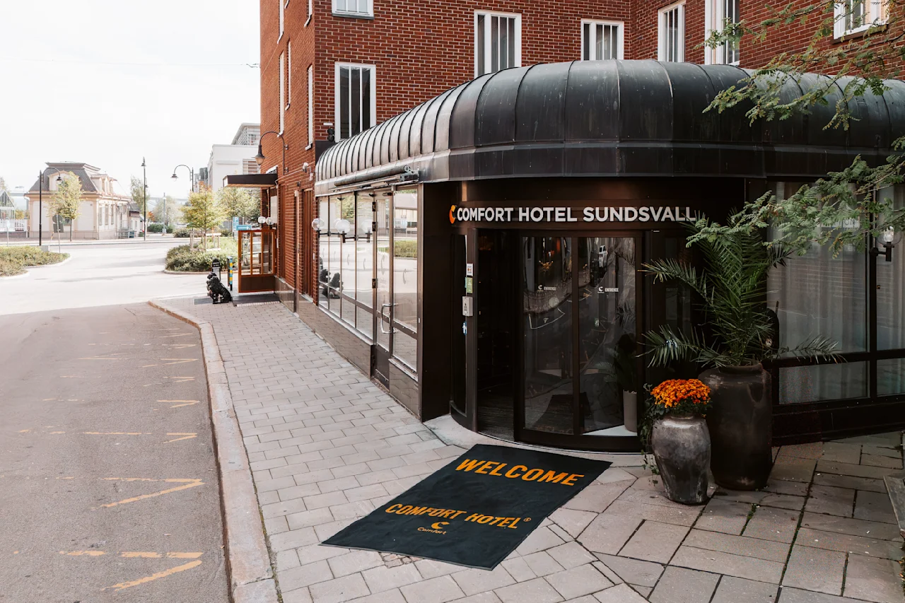 Inngang til Comfort Hotel Sundsvall med sort markise og potteplanter; en velkomstmatte har påskriften "WELCOME COMFORT HOTEL."