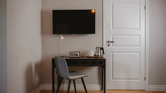 Ett litet skrivbord med stol har en lampa, en vattenkokare och en radio under en väggmonterad TV. Det står i ett rum med trägolv och en stängd vit dörr.