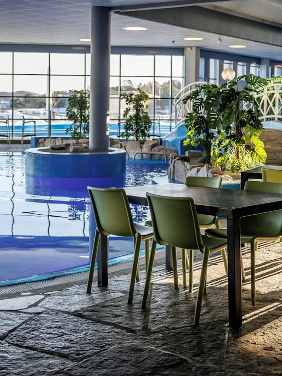 Et basseng med klart vann reflekterer sollys innendørs. I nærheten står bord med grønne stoler på et steingulv. Grønne planter og en hvit dekorativ bro forbedrer stemningen.