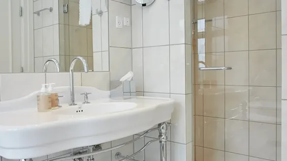 Et lyst, moderne badeværelse med en hvid piedestalvask, kromarmaturer, et stort spejl og en brusekabine i glas.