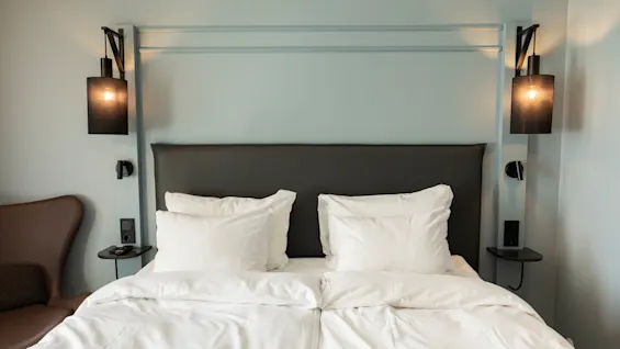 Et koselig hotellrom med en seng, hvitt sengetøy, en mørk sengegavl og veggmonterte lamper på lyseblå vegger.