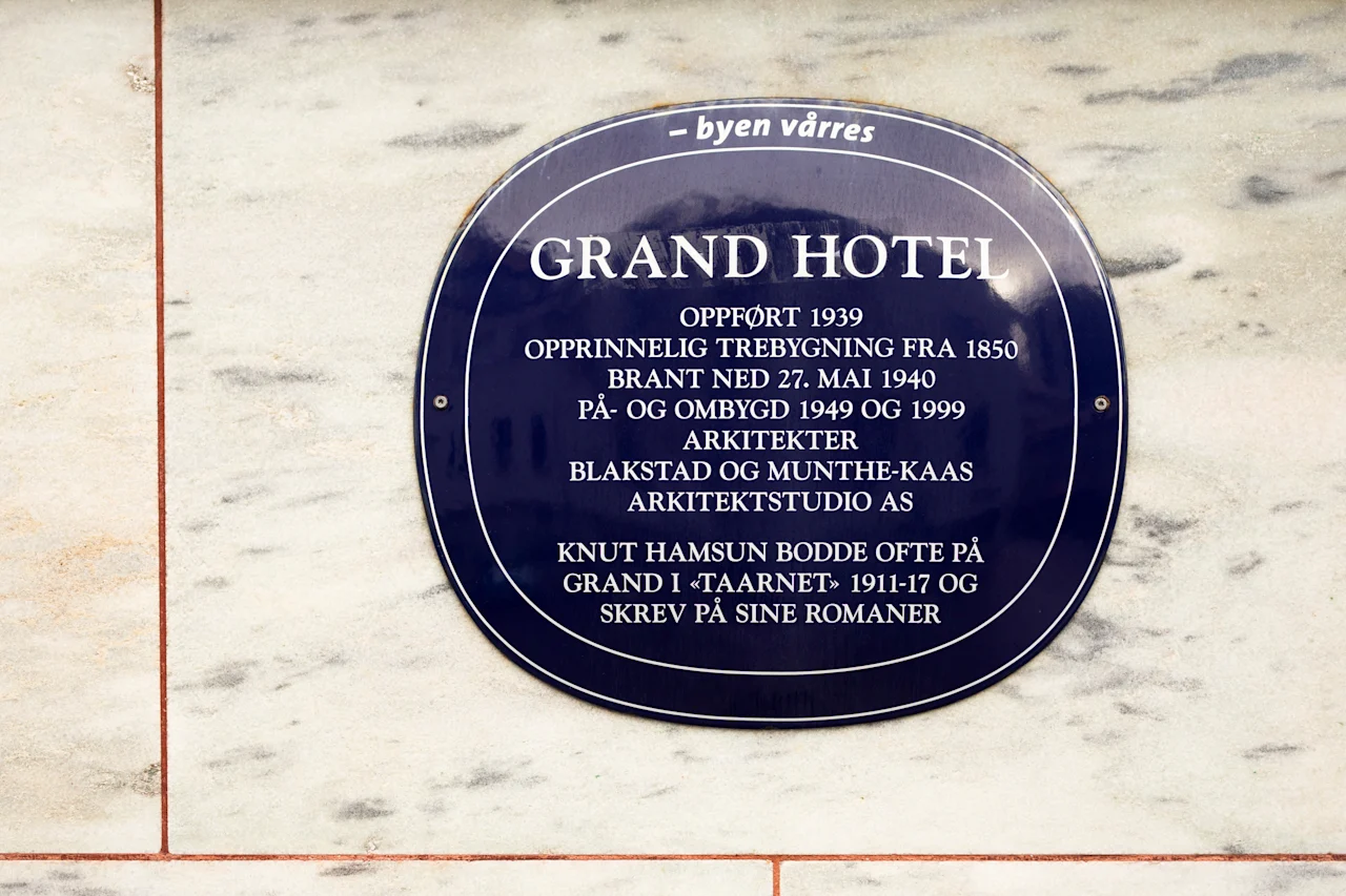 A historical plaque for Home Hotel Grand Bodø, detailing its construction, a fire, renovations, and a famous author's stay.