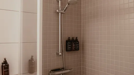Showerhead mounted on a tiled wall, with bottles of shampoo and soap resting on a shelf; adjacent wall features additional toiletries. Warm lighting enhances the clean, minimalist bathroom design.
