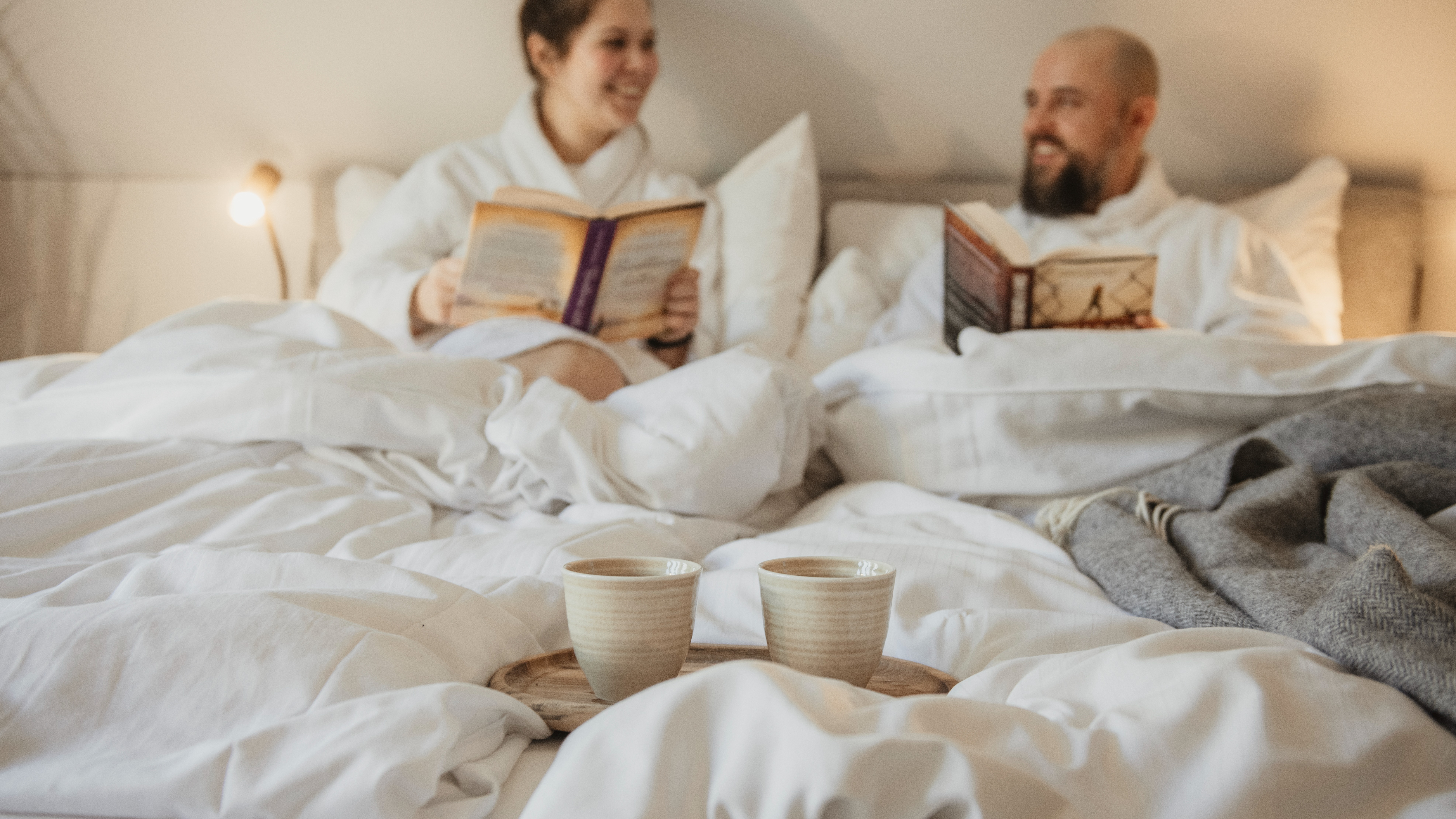 Kaksi ihmistä kylpytakeissa rentoutumassa sängyssä, lukemassa kirjoja, kaksi kuppia kahvia tai teetä tarjottimella etualalla.