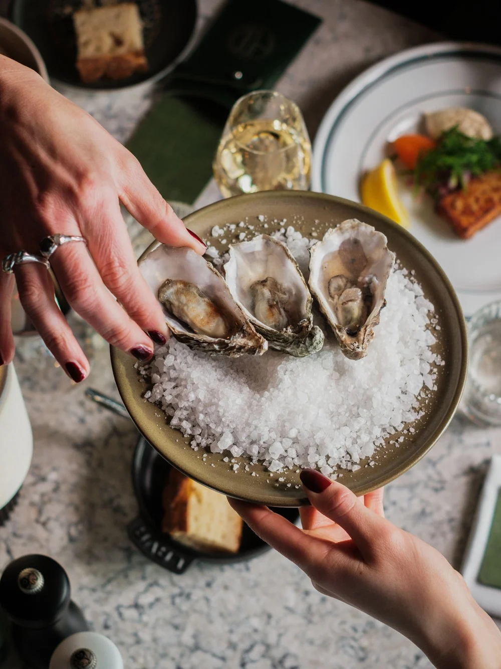 En tallerken med tre østers på knust is overrekkes over et bord. Ulike retter, glass og bestikk omgir det, og skaper en middagssetting.