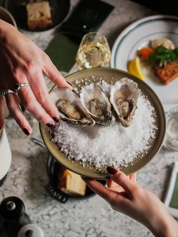 En tallerken med tre østers på knust is overrekkes over et bord. Ulike retter, glass og bestikk omgir det, og skaper en middagssetting.