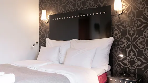 Viihtyisä hotellihuone, jossa on sänky valkoisilla liinavaatteilla, tumma kuvioitu sängynpääty ja lämmin, kutsuva valaistus.