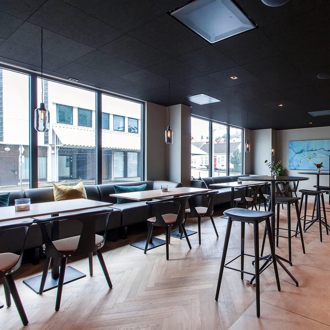 Moderne caféinteriør med stilige bord og stoler og anhengslamper, omgitt av store vinduer og en elegant bardisk. En tavle står: "EXQUISITE" og lister menyartikler. 