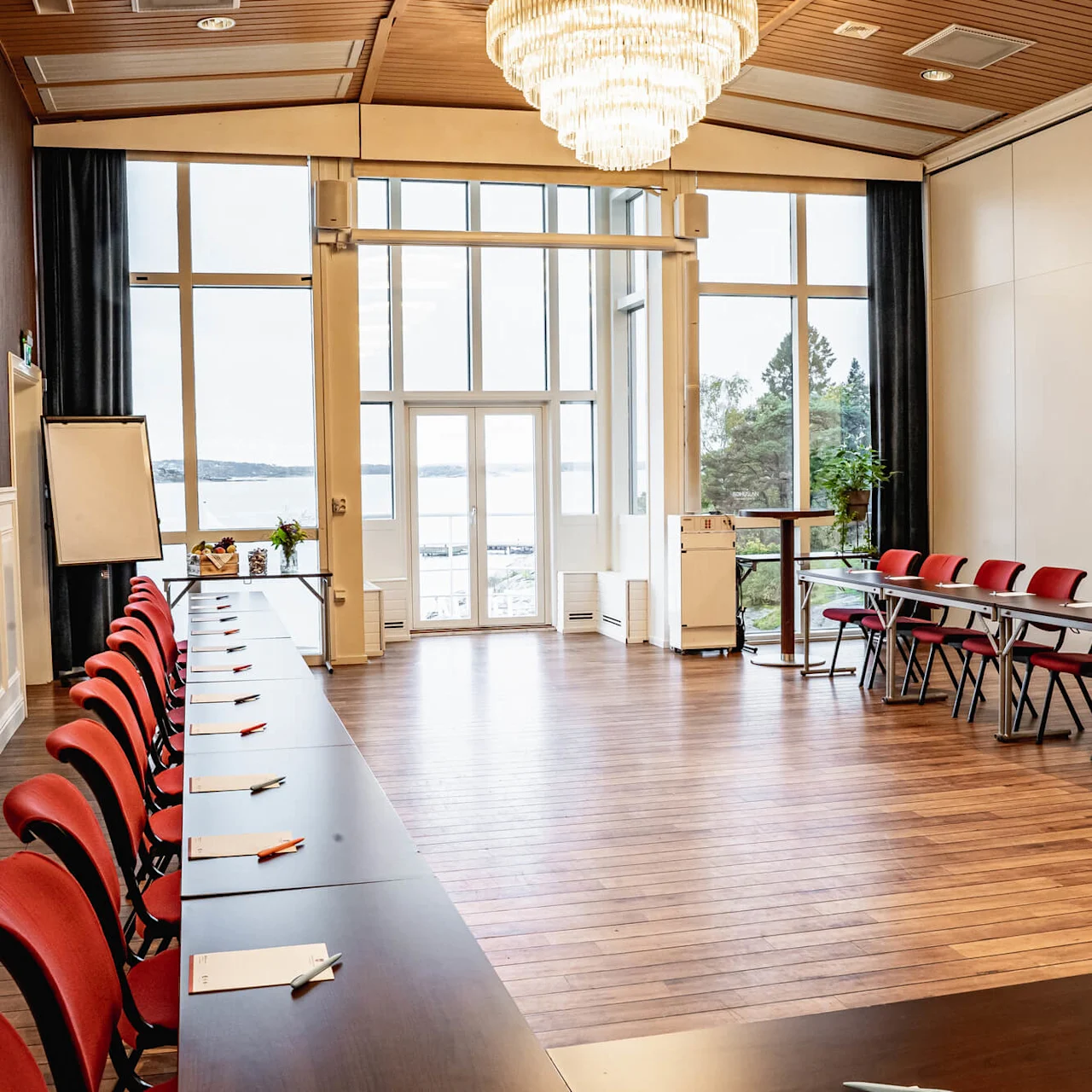 Conference room with red chairs and tables set up facing each other, ready for a meeting. Large windows reveal an outdoor view of water and trees. Papers and pens are arranged on the tables.