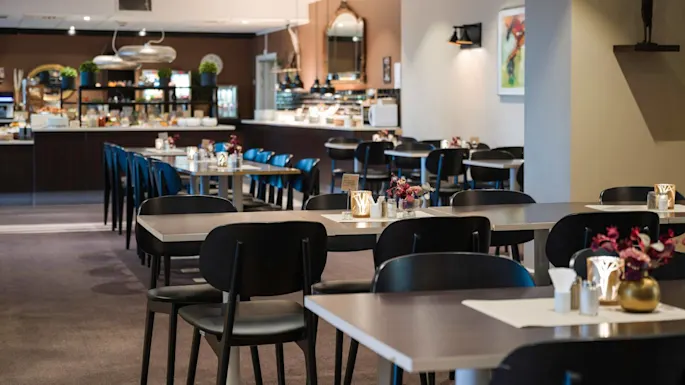 Tables set in a restaurant, adorned with vases and candles, surrounded by chairs. A buffet is visible in the background with lights and decor enhancing the elegant dining atmosphere.