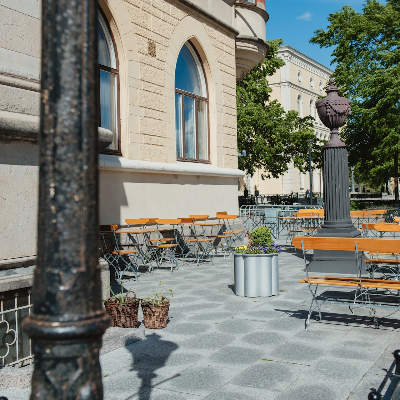 Utomhuscafébord och stolar ordnade på en solbelyst stenlagd uteplats bredvid en historisk byggnad, omgiven av krukväxter och träd under en klarblå himmel.