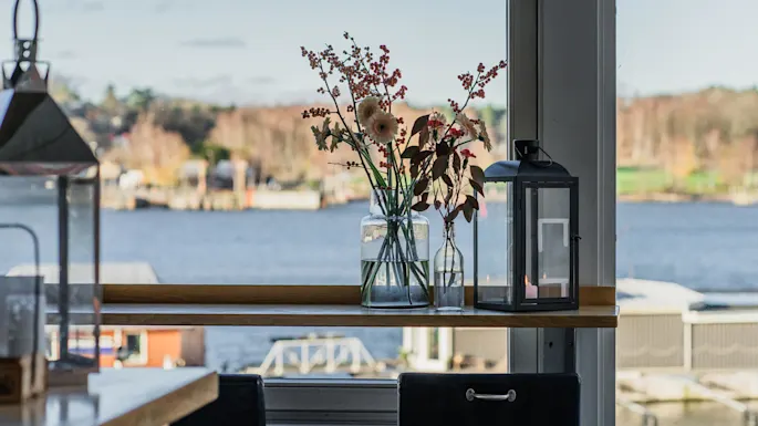En glasvase med blomster og en sort lanterne står på en vindueskarm med udsigt til roligt vand og slørede træer i baggrunden.